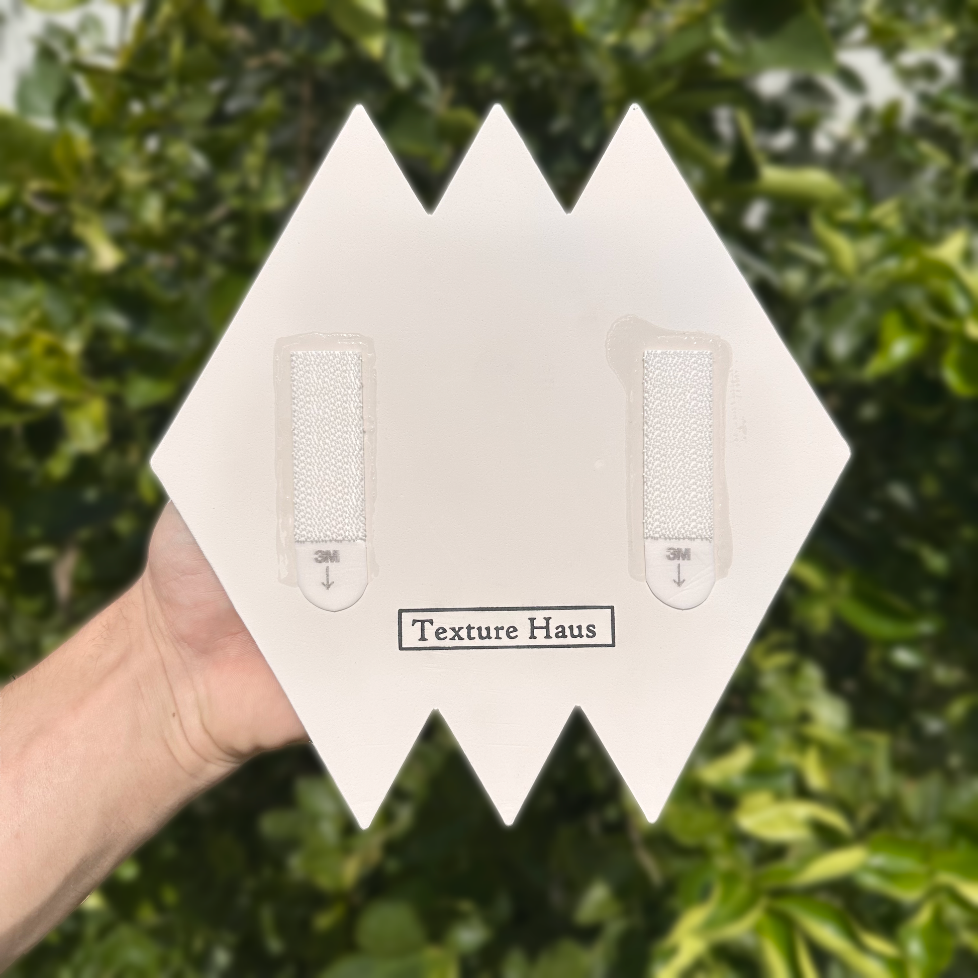 Hand holding a textured wall panel with 'Texture Haus' branding against a green leafy background